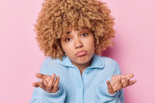 Horizontal shot of curly haired woman spreads palms feels hesitant expresses uncertainty purses lips dressed in blue jumper feels puzzled isolated over pink background. Who knows I dont care