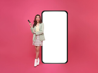 Happy asian woman using mobile phone standing at big giant smart phone with empty white screen,...