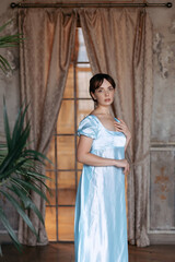 A young beautiful girl in a blue Regency dress stands at a wooden window in the estate. The classic artistic image of Russian literature of the 19th century, outdated.
