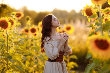 A beautiful young tall slender brunette woman in a beige dress and leather corset stands in a field with blooming yellow sunflowers at sunset. 