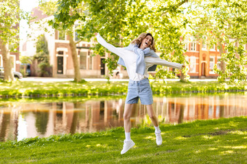Fototapeta premium Portrait of beautiful woman smiling and jumping in park at sunny day. Outdoor portrait of a smiling curly blonde girl. Happy cheerful girl laughing at park wear sweater, white long sleeves, shorts