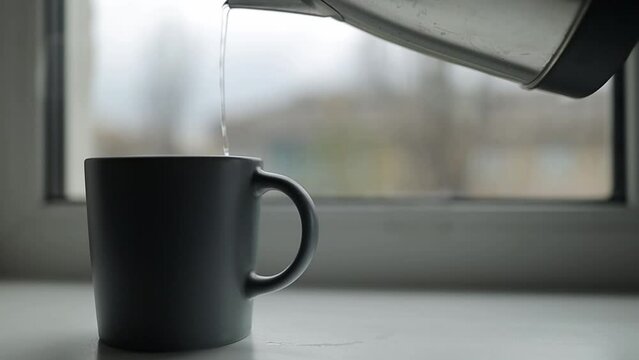pours boiling water from an electric kettle into coffee in a gray mug. pour hot boiled water from an electric kettle into a mug. The kettle pours boiling water into a cup on the windowsill.