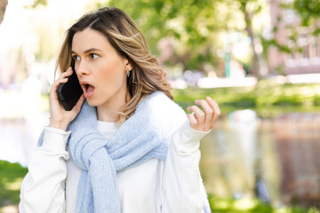 Portrait worried young woman with surprised expression talking on mobile phone, unpleasant call outdoors. Shocked woman calling on smartphone with open mouth and eyes, look at side, spread hand.