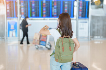 Young woman with bag and luggage checking flight time information board in international airport,...