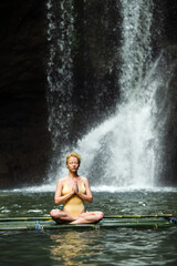 Fototapeta na wymiar The girl is meditating, her hands are folded in namaste, in the jungle against the backdrop of a waterfall, dressed in a yellow swimsuit.