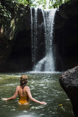 A young sexy girl in a yellow swimsuit bathes in a river with a waterfall in Bali.