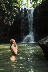 The girl is meditating, her hands are folded in namaste, in the jungle against the backdrop of a waterfall, dressed in a yellow swimsuit.