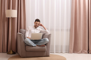 Smiling man holding laptop on armchair near window with beautiful curtains at home. Space for text