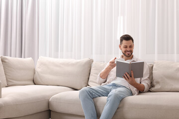 Obraz premium Happy man drinking coffee while reading book on sofa near window with beautiful curtains at home in living room