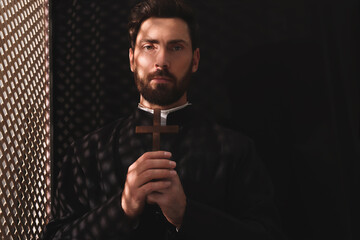 Catholic priest in cassock holding cross in confessional - obrazy, fototapety, plakaty