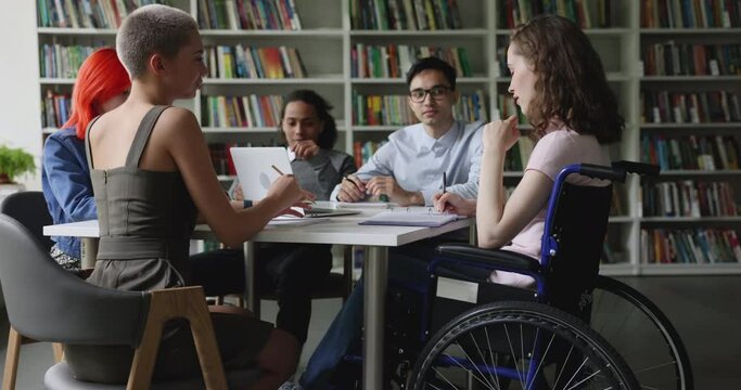 Teen schoolgirl sits in wheelchair take part in group meeting with multi ethnic groupmates discuss joint assignment gathered in library. Accessibility of education of person with physical disability