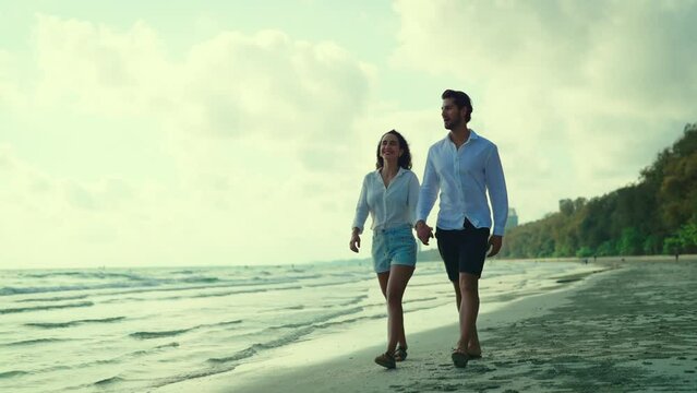 Young couple takes a leisurely stroll along the beach, their smiles beaming as they chat and laugh together, enjoying each other's company in the peaceful setting. Slow motion shot.