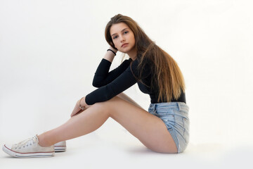 Young long legged girl is sitting on floor on the light background. The girl is looking at the camera.