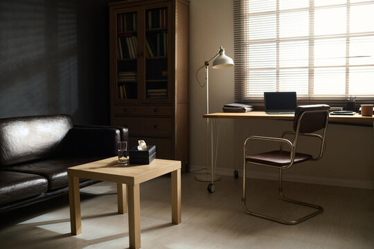 Workplace of psychologist by window and small square wooden table by black comfortable leather couch standing by wall in psychotherapy office