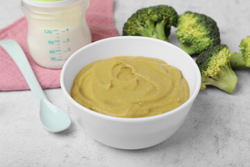 Bowl with healthy baby food, broccoli, spoon and bottle of milk on white table, closeup