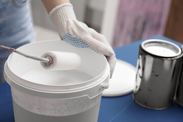 Woman dipping roller into bucket with white paint at blue wooden table indoors, closeup. Space for...