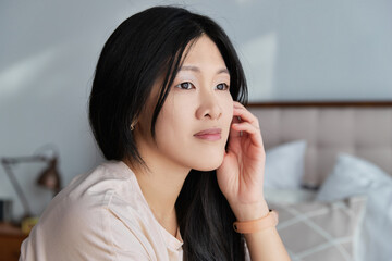 Close up serious asian woman in casual clothes looking out the window sitting on the bed at home