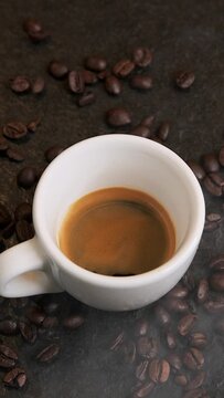 Vertikale Nahaufnahme eines köstlichen frischen Espresso in weißer Tasse vor schwarzem Hintergrund mit frisch gerösteten Espressobohnen die in Slowmotion ins Bild springen