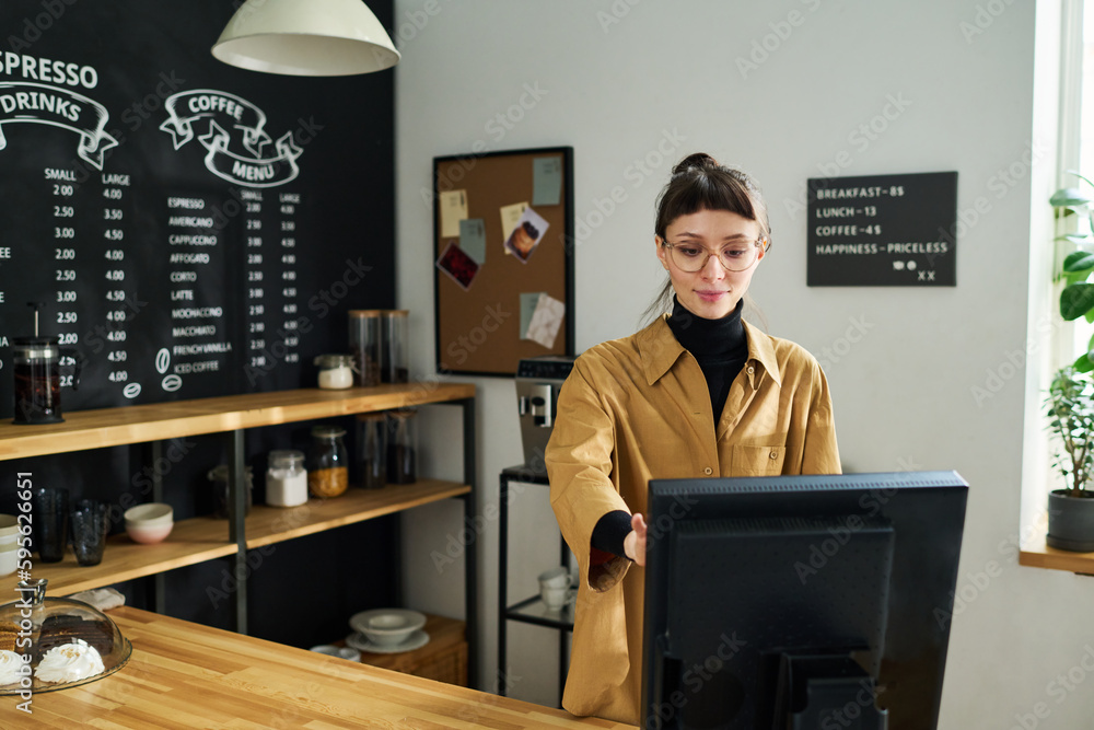 Poster young pretty woman in casualwear and eyeglasses standing in front of computer by counter desk and co