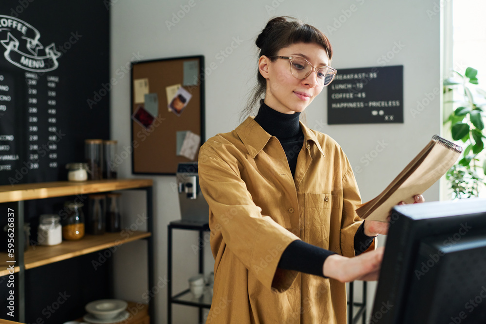 Poster Young pretty waitress with notepad counting price of client order while standing by workplace in cozy cafe and entering data in computer