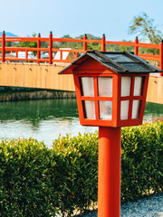 Beautiful view of outdoor lamps on walkway for garden lighting in summer park with blurred image of red bridge in background. Pathway in park with the light pole. Red wooden lantern on pole.