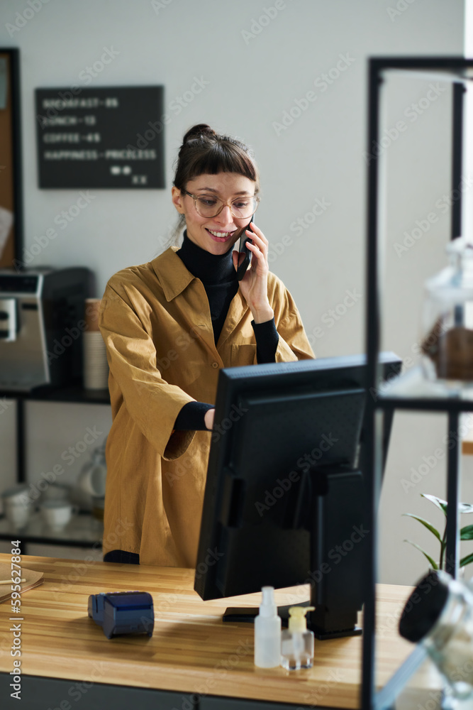 Poster Happy young clerk with smartphone by ear taking order of client and entering data in computer while standing by counter desk in cafe