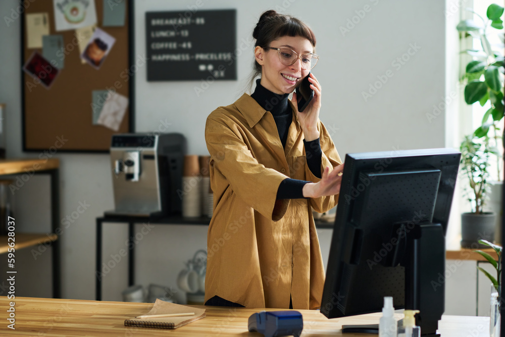 Poster Young smiling female clerk talking with client on smartphone and entering order information in computer while standing by counter desk