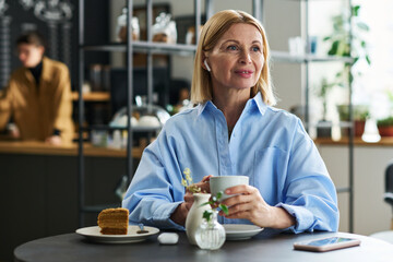 Happy blond mature woman with earphones listening to music while sitting by table in cafe and having cup of tea or coffee with cake