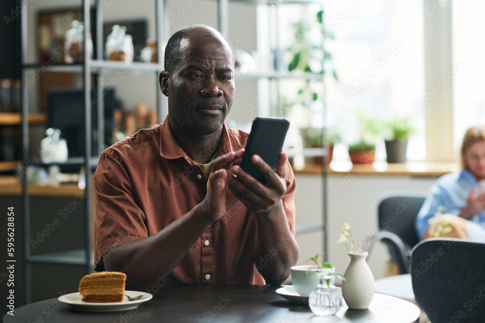 Poster serious mature man with mobile phone texting or looking through online news while sitting by table i