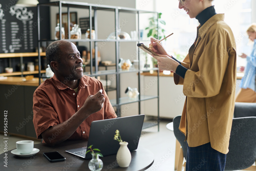 Poster Side view of young waitress in uniform writing down order of happy African American mature client in notepad while standing in front of him