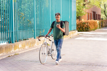 Fototapeta premium Cheerful ethnic man with bicycle walking with backpack delivering food
