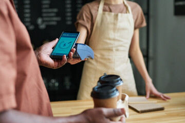 Focus on smartphone held by mature African American male consumer during contactless payment for coffee by counter desk in cafe