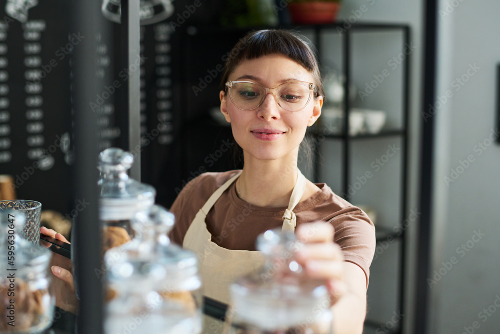 Poster young smiling female clerk checking leftover of dry herbal tea in small jars standing on shelf while