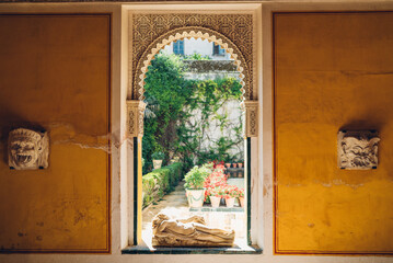 Fototapeta premium Big window frame with a naked woman statue showing a sunny garden in the Casa de Pilatos palace with masks on the yellow walls.
