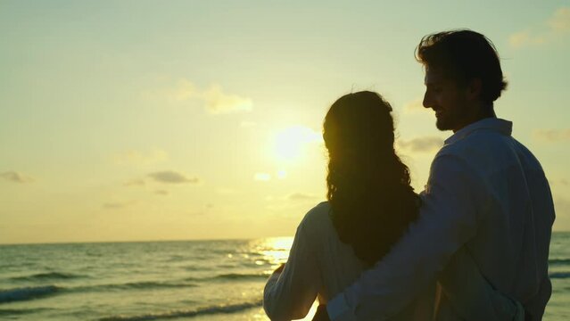 Standing side by side, a young couple gazes out at the sea with a deep sense of romance, their eyes fixed on the stunning sunset that paints the sky with a warm, orange glow. Slow motion shot.
