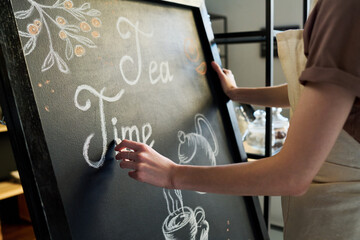 Close-up of young female worker or owner of cafe drawing letters with chalk on big blackboard while...