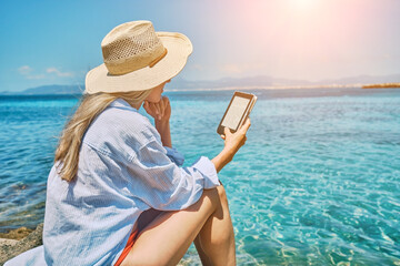 Woman relaxing outdoors, watching, reading on tablet ebook on the beach in summer day. Wearing wide brimmed hat.