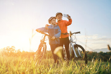 Mother and son ride a bike. Happy Family with mobile phone, cute boy in helmet learn to riding a bike in park on green meadow at sunset time. Family weekend.
