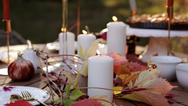 Beautiful Thanksgiving festive fall table setting with white burning candles and autumn leaves close up. Blurred background with eating child and homemade pie on stand. Family celebration dinner.