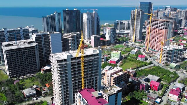 Drone view of construction of high-rise buildings on summer day against backdrop of sea and mountains. Busy construction site and construction equipment on sea coast of Batumi in Georgia.