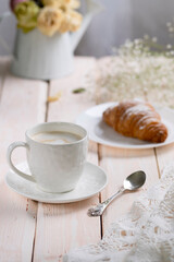 Cappuccino coffee in a white cup with a croissant on a wooden table, high key