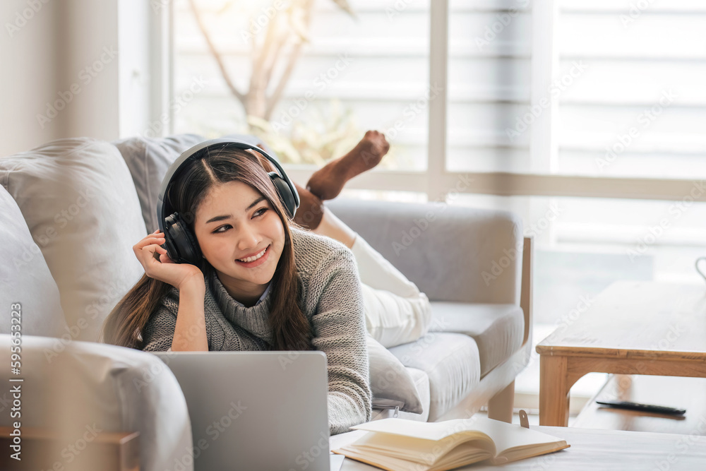 Wall mural Photo of modern asian woman wearing headphones use laptop while lying on sofa in bright home