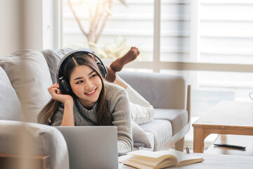 Photo of modern asian woman wearing headphones use laptop while lying on sofa in bright home