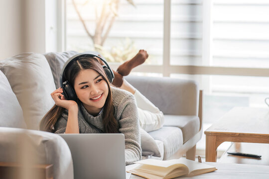 Photo of modern asian woman wearing headphones use laptop while lying on sofa in bright home