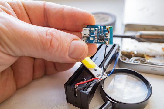 man 's hand holds an electric battery charger board next to the included soldering iron and a tape with a magnifying glass . the concept of repair and maintenance