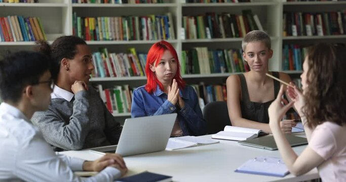 Teen student girl with dyed red hair make speech at group meeting in library with teammates, share ideas, prepare common assignment or project engaged in creative teamwork studying together. Education
