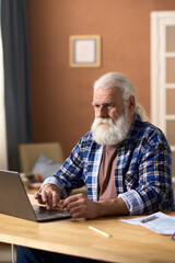 Vertical image of elderly man concentrating on online work on laptop while sitting at table in the living room