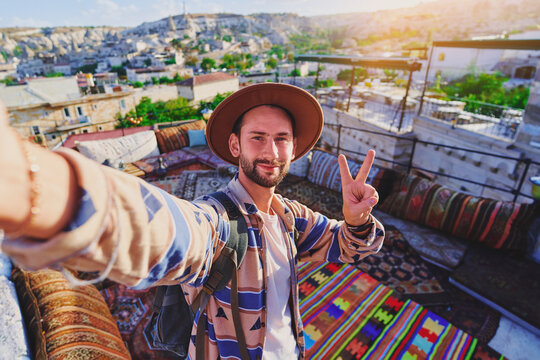 Bearded guy traveling blogger with victory sign takes selfie photo at hotel terrace with colorful carpets in Nevsehir, Goreme. Beautiful destination in Anatolia, Kapadokya