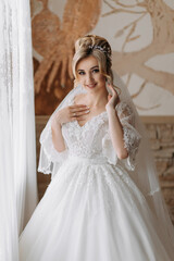 Fototapeta premium A blonde bride in a white dress and a long veil is posing by the window. Beautiful hands. French manicure. Beautiful hair and make-up. Diadem. Daylight