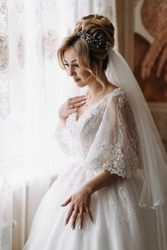 A blonde bride in a white dress and a long veil is posing by the window. Beautiful hands. French manicure. Beautiful hair and make-up. Diadem. Daylight
